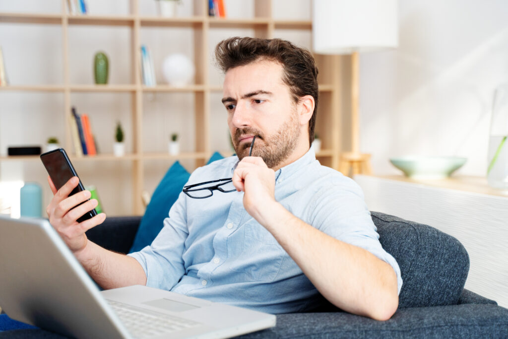 man in front of phone and computer chewing his glasses wondering if no credit check loans are safe or scams