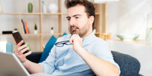 man in front of phone and computer chewing his glasses wondering if no credit check loans are safe or scams